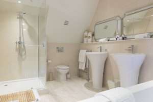 Well-lit en-suite bathroom at Greenwoods Hotel Attic Suite with walk-in glass shower, double basins, and elegant neutral tones.