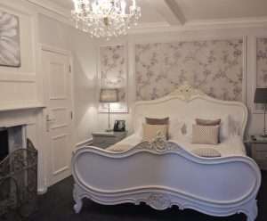 Charming and calming bedroom at Greenwoods Hotel with floral lavender accents, chandelier, and carved cream bed frame.
