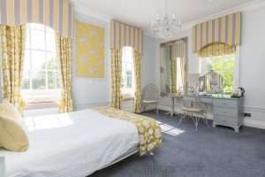 Light-filled guest room at Greenwoods Hotel decorated in yellow tones with elegant mirrors and classic dressing table.