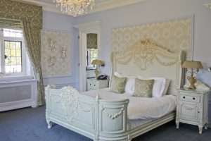 Regal French-inspired bedroom at Greenwoods Hotel with carved white headboard, pastel wallpaper and chandelier lighting.