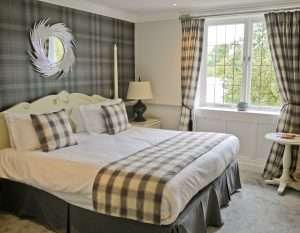 Charming Greenwoods Hotel standard bedroom with matching plaid pillows and curtains, warm lighting, and decorative round wall mirror.
