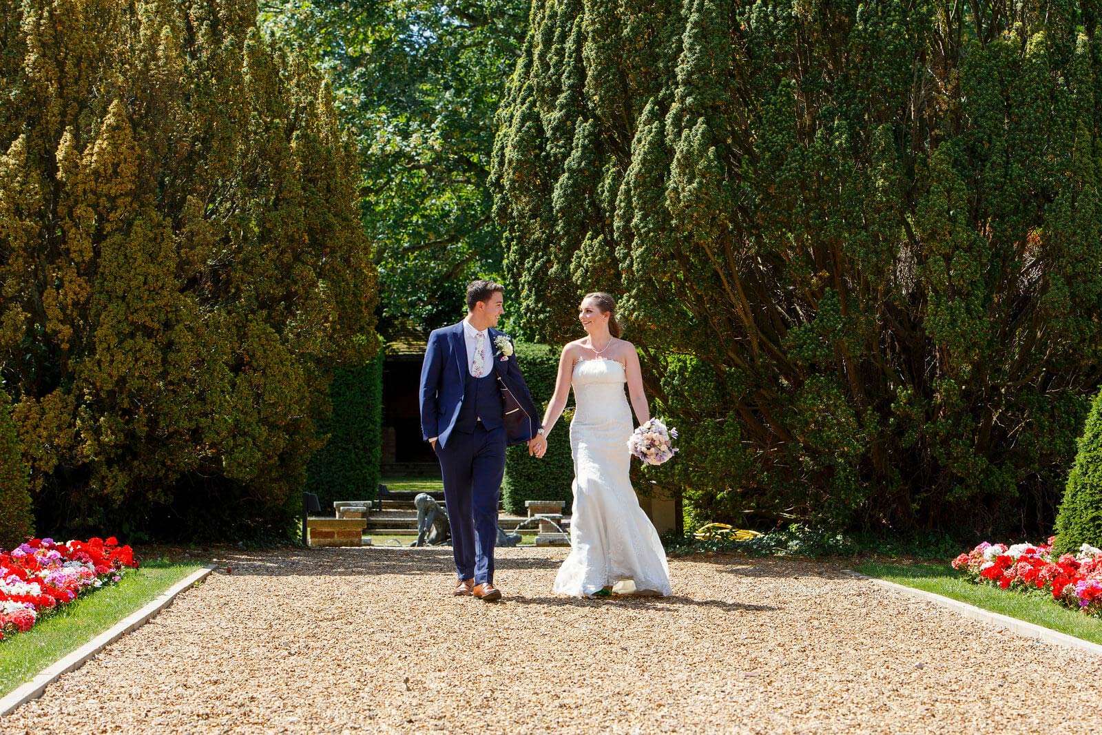 Bride and groom holding hands while walking along the gravel path lined with flowers at Greenwoods Hotel
