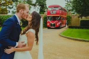 Bride and groom kiss with classic red London wedding bus parked behind at Greenwoods Hotel