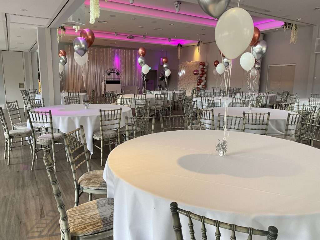 Round banquet tables decorated with clusters of pink, silver, and white balloons for a school prom event.
