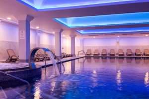 Wide view of the luxury indoor spa pool at Greenwoods Hotel & Spa featuring hydrotherapy water features, ambient blue lighting, and relaxation loungers
