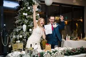 Bride and groom raising champagne during a festive wedding toast beside a Christmas tree at Greenwoods Hotel & Spa in Essex.