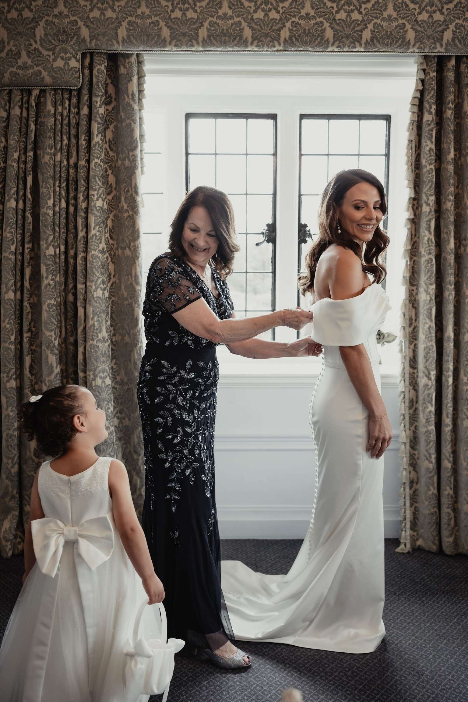 Bride having her wedding dress fastened in a bridal suite at Greenwoods Hotel and Spa, with a young flower girl watching.