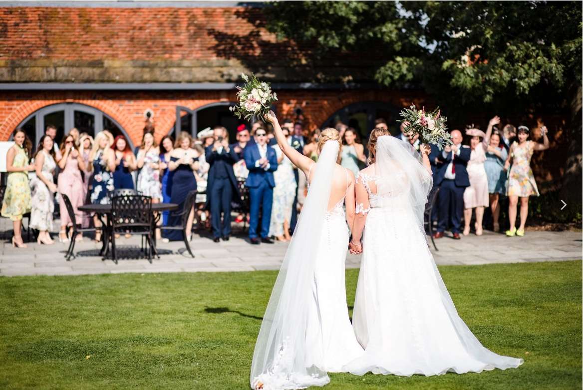 Two brides holding hands and raising bouquets during a garden wedding celebration at Greenwoods Hotel and Spa in Essex.